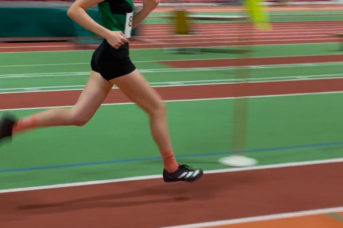 Junge Athletin sprintet auf einer leichtathletischen Bahn, im Hintergrund verschwommene Trib&uuml;nen und Hinweisschilder.