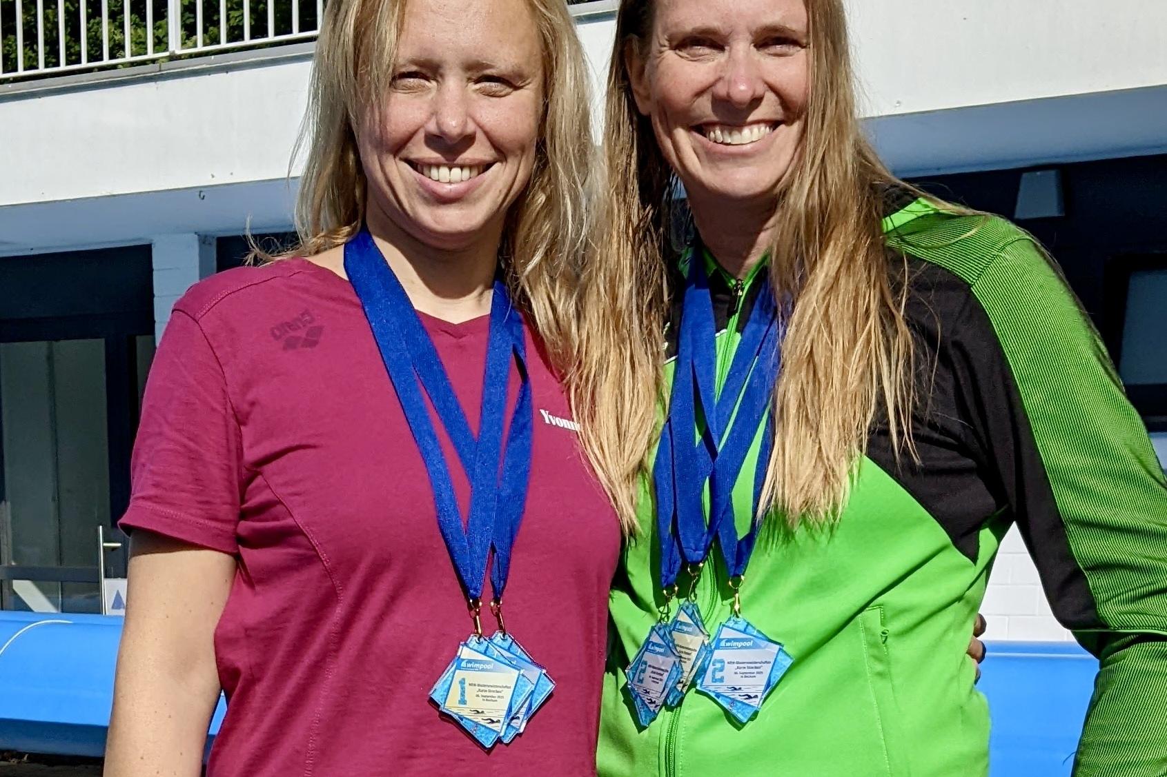 Zwei Frauen stehen nebeneinander und tragen mehrere Medaillen um den Hals, vor einem Schwimmbad im Sonnenlicht.