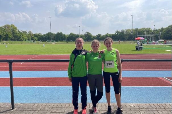 Drei Athletinnen in Sportkleidung posieren auf einem Leichtathletikgel&auml;nde mit gr&uuml;ner Wiese und Laufbahn im Hintergrund.