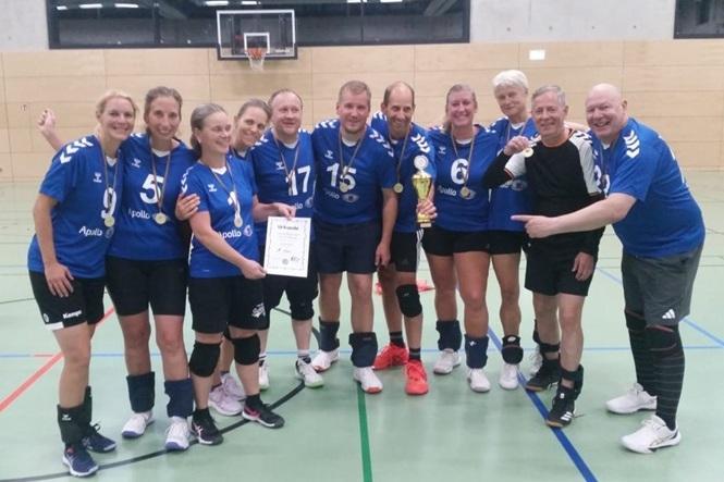Gruppenfoto eines Handballteams in blauen Trikots mit Medaillen, Pokal und Urkunde in einer Sporthalle.