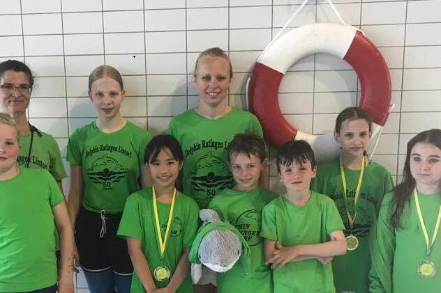 Gruppe von acht Schwimmern in gr&uuml;nen T-Shirts, zwei halten Medaillen, vor einem Schwimmring an einer Wand.