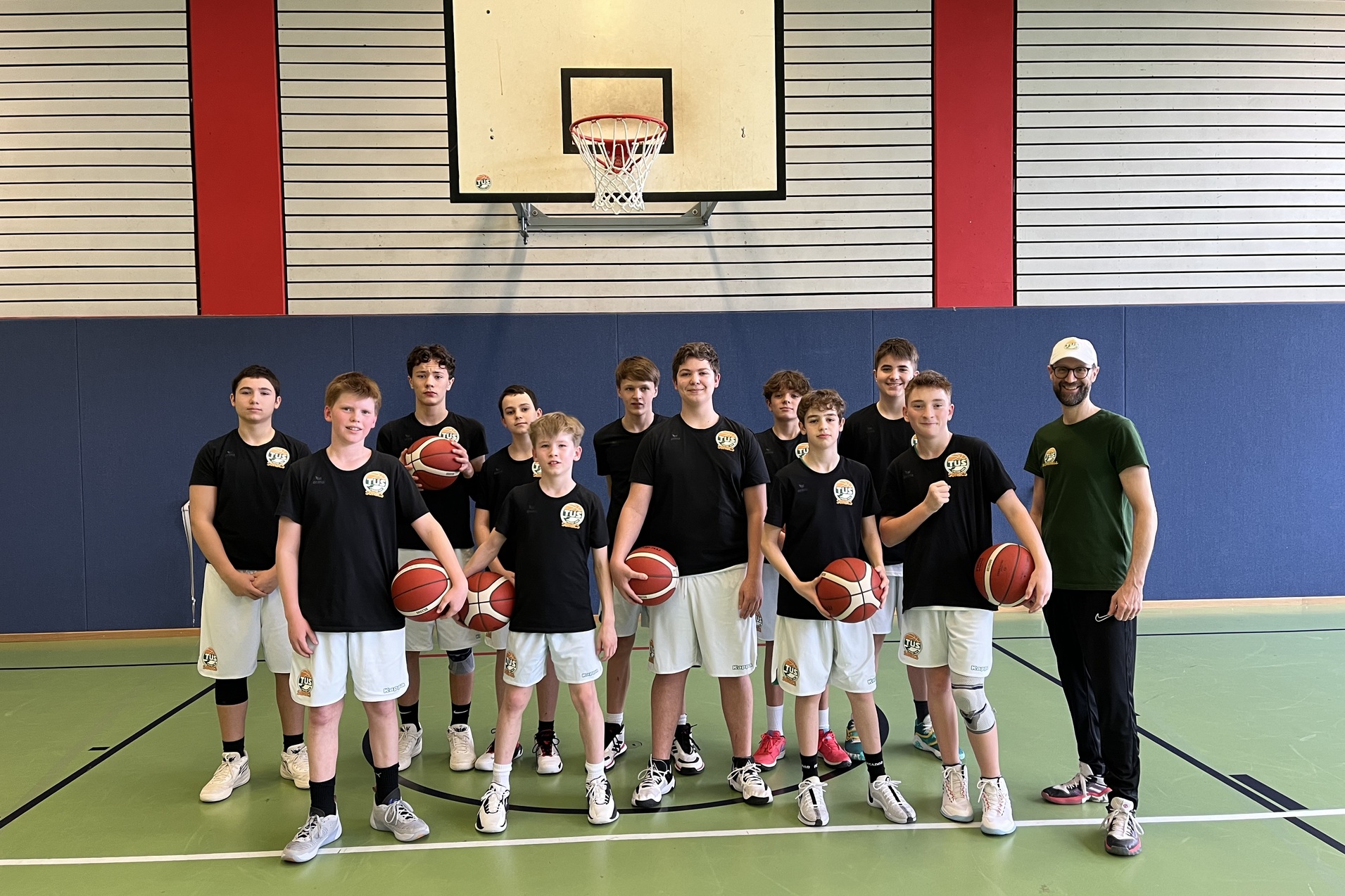 Gruppe von 12 Basketballspielern in schwarzen Trikots mit B&auml;llen auf einem Basketballfeld hinter einem Korb.