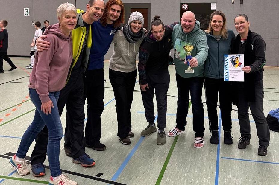 Gruppe von sieben Menschen in Sportkleidung hält einen Pokal und eine Urkunde in einer Turnhalle.