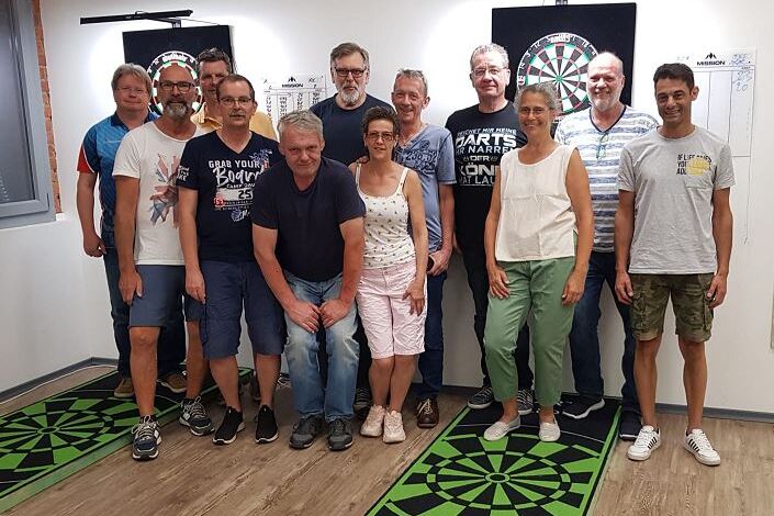 Gruppenfoto von 14 Personen im Darts-Raum mit zwei Dartboards und gr&uuml;nen Dartscheiben-Design-Matten.