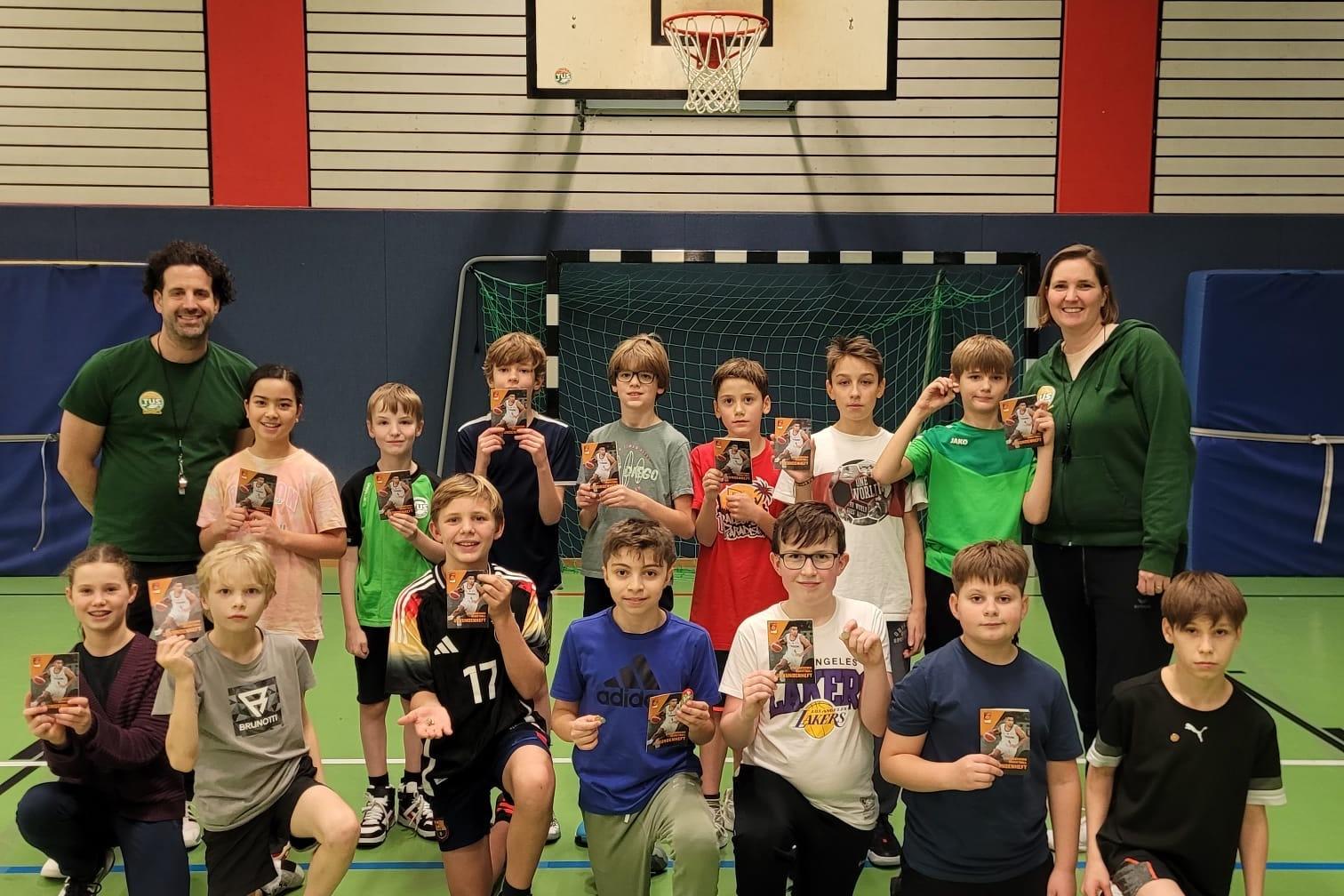 Gruppe von Kindern und Betreuern hält Pokale in einer Sporthalle mit Basketballkorb im Hintergrund.