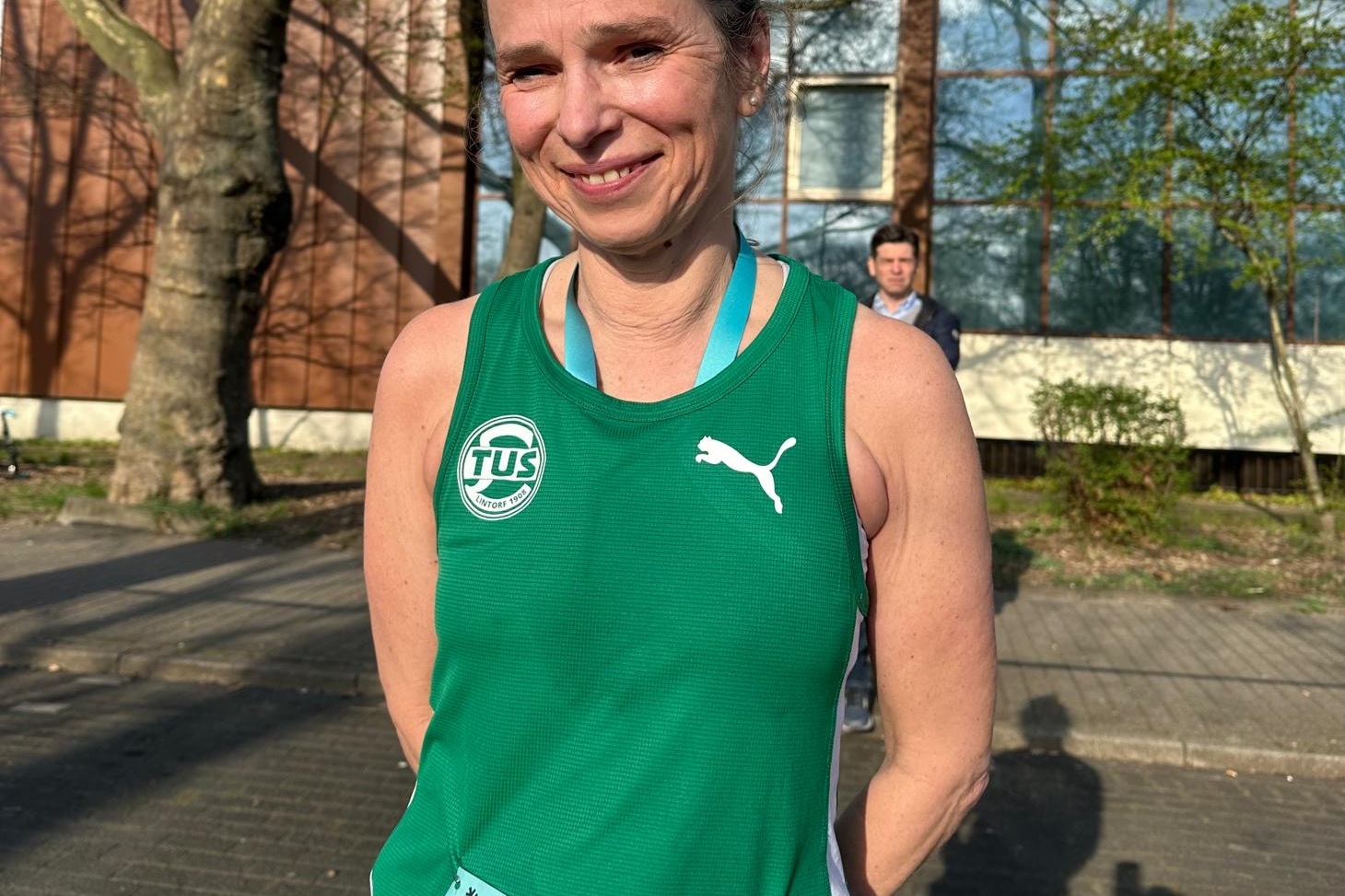 Frau in gr&uuml;ner Laufbekleidung mit Startnummer F764 steht vor einem Geb&auml;ude und l&auml;chelt.