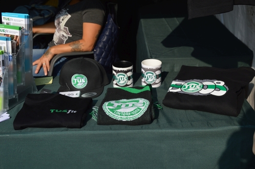 Tisch mit verschiedenen Merchandise-Artikeln, darunter T-Shirts, Caps und Tassen, alle mit dem Logo &bdquo;TU S&ldquo;.