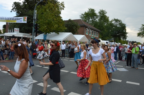 Tanzende Menschen in bunten Kleidern auf einer Stra&szlig;e, umgeben von Zuschauern und Marktst&auml;nden.