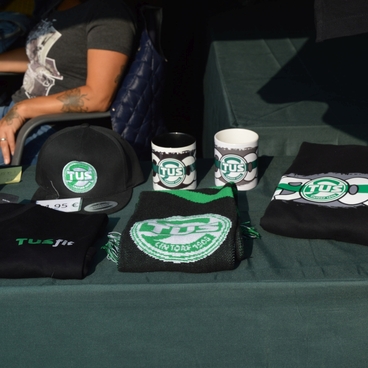Tisch mit verschiedenen Merchandise-Artikeln, darunter T-Shirts, Caps und Tassen, alle mit dem Logo &bdquo;TU S&ldquo;.