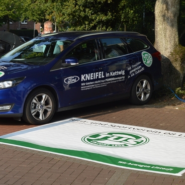 Blauer Ford mit Werbung f&uuml;r "Kneifel in Kettwig" parkt neben einem wei&szlig;en Banner des TUS-Zentrums auf einem gepflasterten Platz.