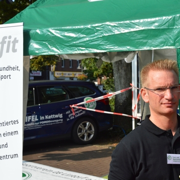 Mann in schwarzem T-Shirt steht vor einem Zelt mit TUSfit-Logo und Informationsbanner &uuml;ber Gesundheit und Fitness.