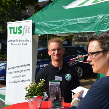 Zwei Personen stehen am Info-Stand von TUSfit, Hintergrund mit Banner und Werbematerial zum Gesundheits- und Fitnessangebot.