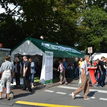 Personen vor einem Informationsstand mit gr&uuml;ner Markise auf einer belebten Stra&szlig;e, umgeben von B&auml;umen.