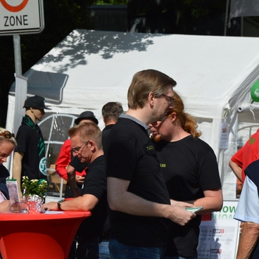 Menschen stehen an einem Stand im Freien, umgeben von Zelten und Informationsmaterialien.