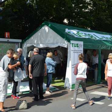 Menschen stehen vor einem Zelt mit der Aufschrift &bdquo;TUSfit&ldquo; an einer Stra&szlig;enecke, umgeben von B&auml;umen und Verkehrssignal.