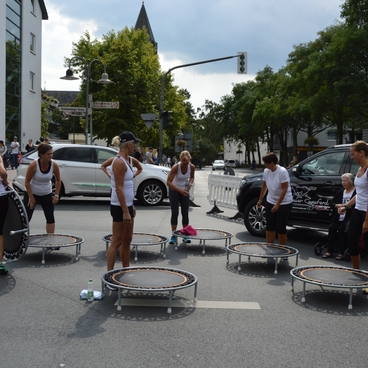 Gruppe von Menschen in Sportbekleidung springt auf Mini-Trampolinen auf einer Stra&szlig;e, umgeben von Autos und Zuschauern.