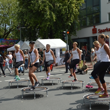 Gruppentraining auf Mini-Trampolinen in der Stadt, umgeben von Zuschauern und Marktst&auml;nden.