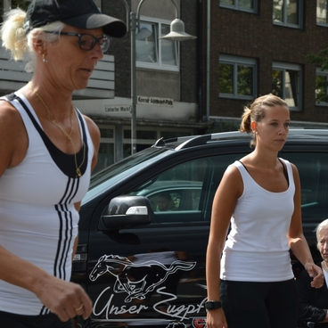 Zwei Frauen in Sportkleidung gehen entlang einer Stra&szlig;e, w&auml;hrend eine &auml;ltere Frau auf einem Stuhl sitzt.