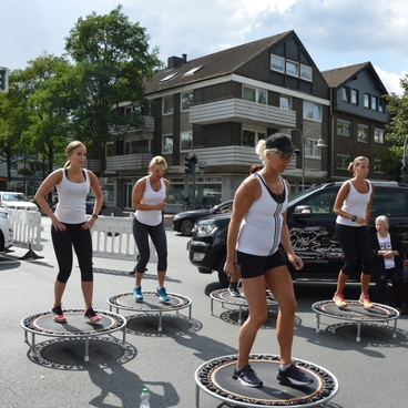 Gruppentraining auf Mini-Trampolinen in einer urbanen Umgebung, mit Frauen in Sportkleidung und einer Zuschauerin.