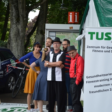 Gruppe von Personen in Freizeitkleidung steht vor einem Zelt mit einem Banner f&uuml;r TUSfit, Zentrum f&uuml;r Gesundheit und Sport.