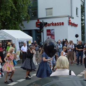 Gruppentanz auf einer Stra&szlig;e vor einer Bank, umgeben von Zuschauern, mit bunten Kost&uuml;men und Luftballons.