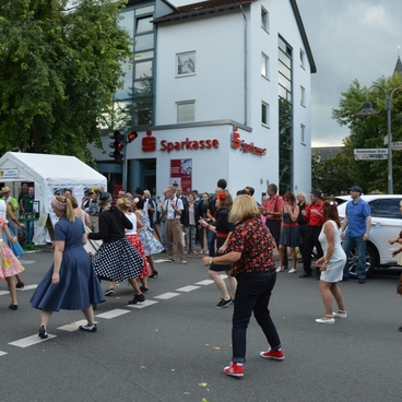Tanzende Menschen in bunter Kleidung auf einer Stra&szlig;e vor einem Sparkassen-Geb&auml;ude, umgeben von Zuschauern und St&auml;nden.