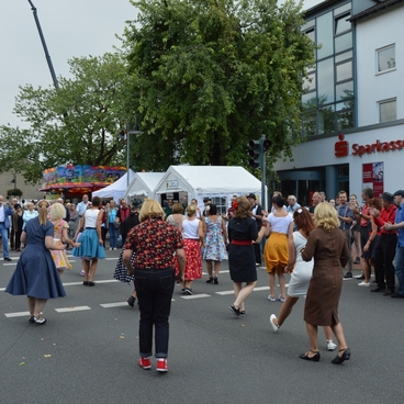 Menschen in festlicher Kleidung tanzen und feiern auf einer Stra&szlig;e vor St&auml;nden und einer Bank.