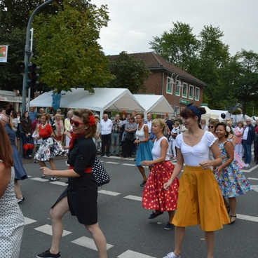 Tanzende Menschen in bunten Kleidern auf einer Stra&szlig;e, umgeben von Zuschauern und Marktst&auml;nden.