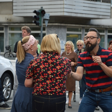 Tanzende Menschen auf einer Stra&szlig;e, umgeben von Zuschauern und Fahrzeugen, bei einem &ouml;ffentlichen Event.