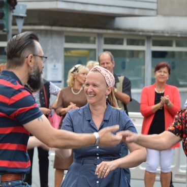 Drei Personen tanzen auf einer Stra&szlig;e, w&auml;hrend eine Gruppe von Zuschauern im Hintergrund applaudiert.