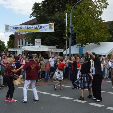 Tanzende Menschen auf einem Handwerker-Markt mit Festzelten und einer bunten Menschenmenge im Hintergrund.
