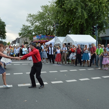 Tanzpaar in festlicher Kleidung f&uuml;hrt einen Lindy-Hop auf einer Stra&szlig;e vor einer Zuschauermenge auf.