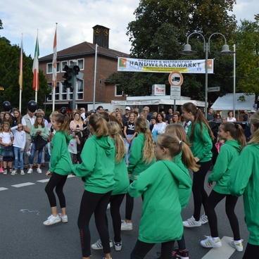 Gruppe von T&auml;nzerinnen in gr&uuml;nen Jacken f&uuml;hrt eine Choreografie vor einer gro&szlig;en Zuschauermenge auf einer Stra&szlig;e auf.