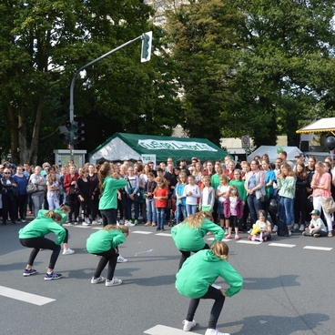 Tanzgruppe in gr&uuml;nen T-Shirts f&uuml;hrt Performance vor einer gro&szlig;en Menschenmenge auf einer Stra&szlig;e durch.