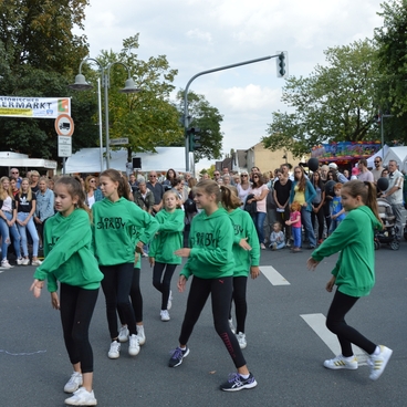 Tanzgruppe in gr&uuml;nen Shirts f&uuml;hrt eine Choreografie vor einer gr&ouml;&szlig;eren Menschenmenge auf einem Stra&szlig;enfest auf.