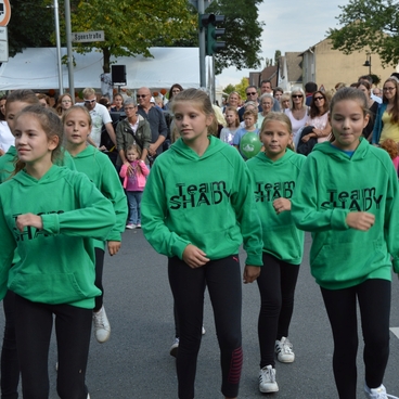 F&uuml;nf M&auml;dchen in gr&uuml;nen Sweatshirts tanzen auf einer Stra&szlig;e vor einem Publikum.
