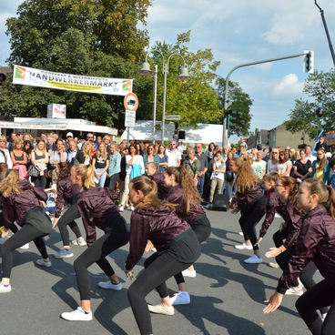 Tanzgruppe in sportlichen Outfits f&uuml;hrt eine Choreografie vor einer gro&szlig;en Menschenmenge auf einem Stra&szlig;enfest vor.