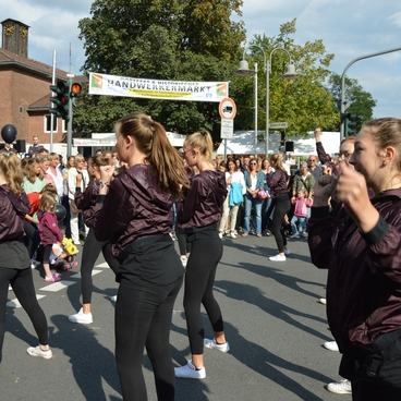 Tanzende Gruppen von Jugendlichen in schwarzen Jacken w&auml;hrend eines Festumzugs auf einer belebten Stra&szlig;e.