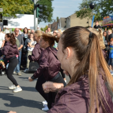 Tanzende Gruppe in lila Jacken vor Publikum auf einer Stra&szlig;e, umgeben von St&auml;nden und festlicher Atmosph&auml;re.