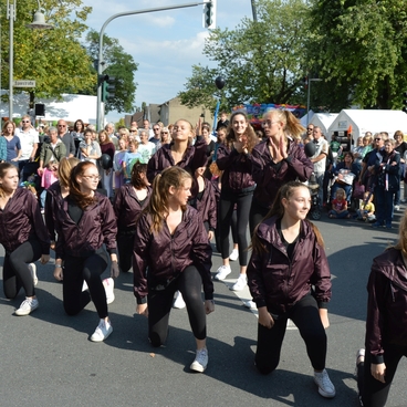 Tanzgruppe in lila Jacken f&uuml;hrt eine Choreografie auf einer belebten Stra&szlig;e vor Publikum auf.