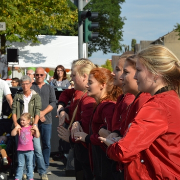 F&uuml;nf junge Frauen in roten Jacken f&uuml;hren eine Tanzperformance vor einem Publikum auf einer belebten Stra&szlig;e durch.