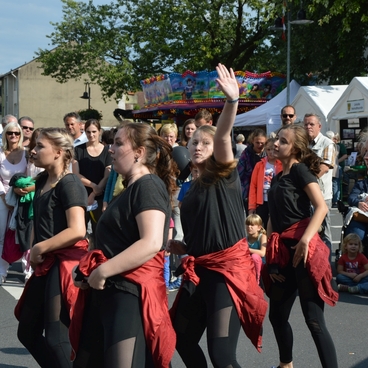 Tanzgruppe in schwarzen Outfits mit roten T&uuml;chern bei einer Auff&uuml;hrung vor einer Zuschauergruppe auf einer Stra&szlig;e.