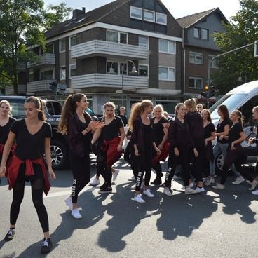 Gruppe junger T&auml;nzerinnen in schwarzen Outfits auf einer Stra&szlig;e, umgeben von parkenden Autos und Stadtgeb&auml;uden.