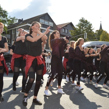 Tanzgruppe in schwarzen Outfits mit roten T&uuml;chern, die auf einer Stra&szlig;e eine Choreografie auff&uuml;hrt. Zuschauer beobachten.