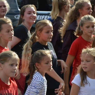 Gruppe von M&auml;dchen in bunten T-Shirts, die gemeinsam an einer Veranstaltung teilnehmen und sich zur Musik bewegen.