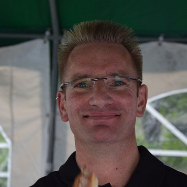 L&auml;chelnder Mann mit Brille h&auml;lt Essen in der Hand, im Hintergrund eine gr&uuml;ne Zeltumgebung.