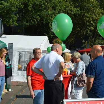 Menschen unterhalten sich auf einem Stra&szlig;enfest, gr&uuml;ne Luftballons schweben &uuml;ber einem Stand und einem Zelt.