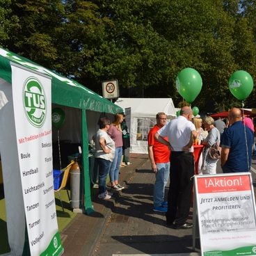 Menschen stehen an einem Stand mit gr&uuml;nen Ballons, vorbei an einer Werbetafel und einem Zelt mit Vereinsinformationen.
