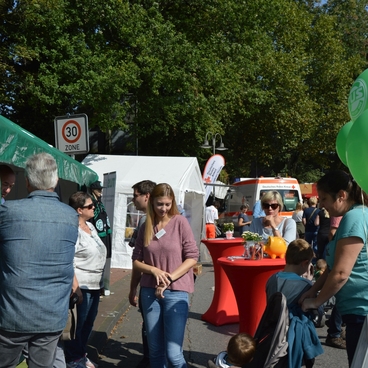 Menschen sind auf einem Stra&szlig;enfest versammelt, umgeben von St&auml;nden und gr&uuml;nen Ballons.