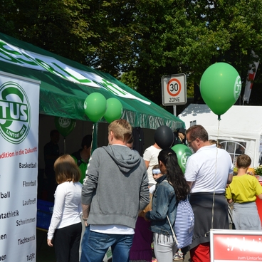 Menschen stehen an einem Informationsstand mit gr&uuml;nen Ballons, um Angebote von TUS zu entdecken.